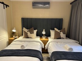 Habitación de hotel con dos camas, una mesita de noche con lámpara, aire acondicionado de pared y una ventana con cortinas.