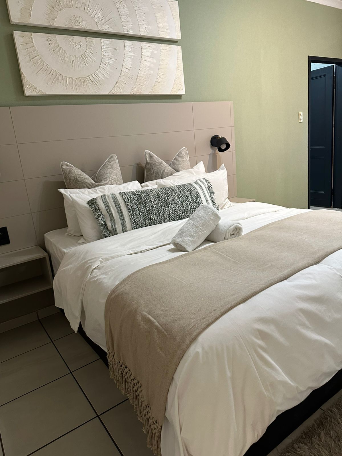 Una cama bien hecha con almohadas y una manta, un cabecero, una lámpara de pared y una puerta.