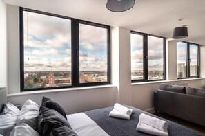 Ein modernes Hotelzimmer mit großen Fenstern und Blick auf die Stadt, einem Bett mit Kissen, einem grauen Sofa und einer Pendelleuchte.