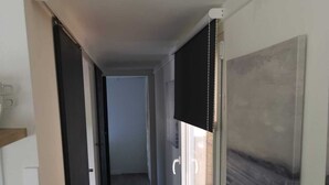 Un pasillo estrecho con piso oscuro, una ventana con persianas y un cuadro en la pared.