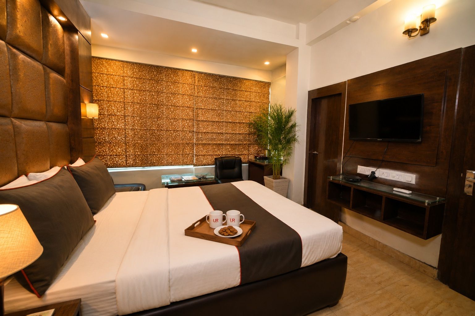 Une chambre d’hôtel moderne avec un grand lit, un téléviseur à écran plat, un bureau avec une chaise, une plante en pot et un plateau avec deux tasses et des biscuits.