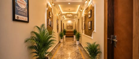 A hotel corridor with a sign indicating mask and social distancing requirements, potted plants, a chandelier, and framed artwork on the walls.