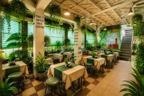 A restaurant with tables and chairs, green plants, and a mural on the wall. - HOTEL UR ( Nearby Prayagraj Junction & High Court) (Prayagraj)