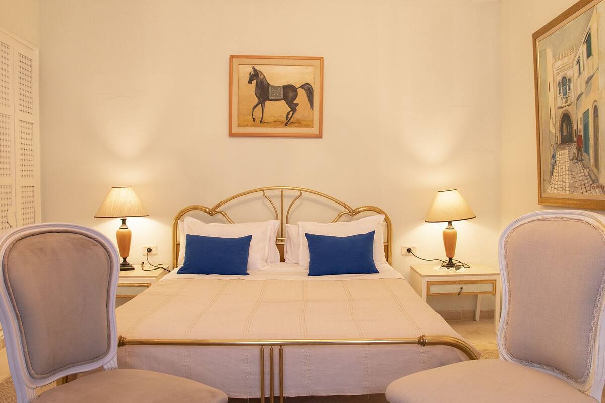 A hotel room with a bed, two chairs, two bedside tables with lamps, a painting on the wall, and a window with a view of buildings.