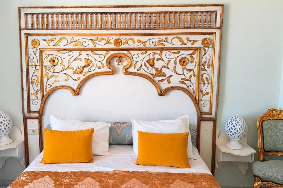 A bed with two white and two orange pillows, a patterned bedspread, and a decorative headboard with a floral design.