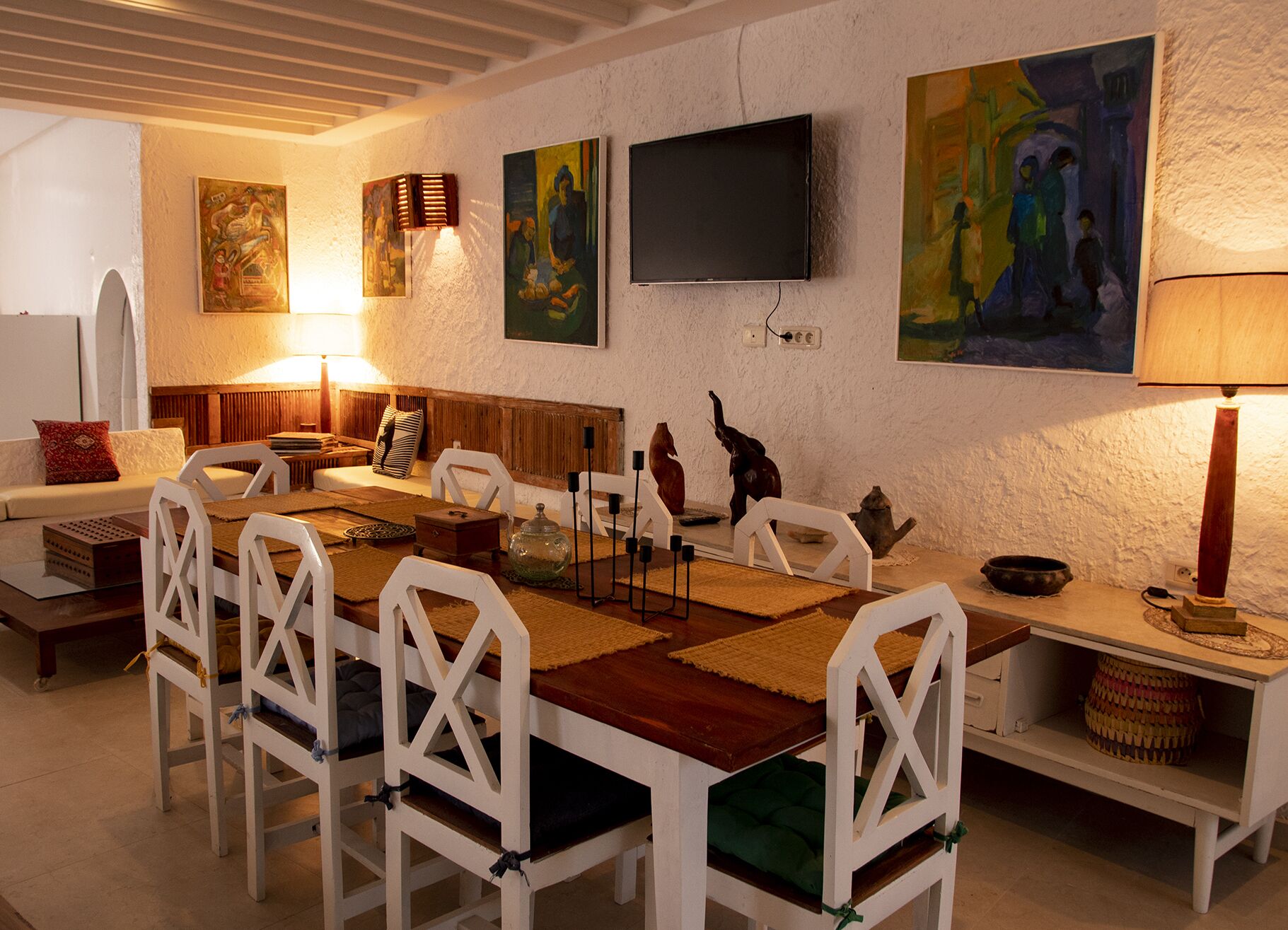 A dining area with a long table, white chairs, and a TV mounted on the wall.