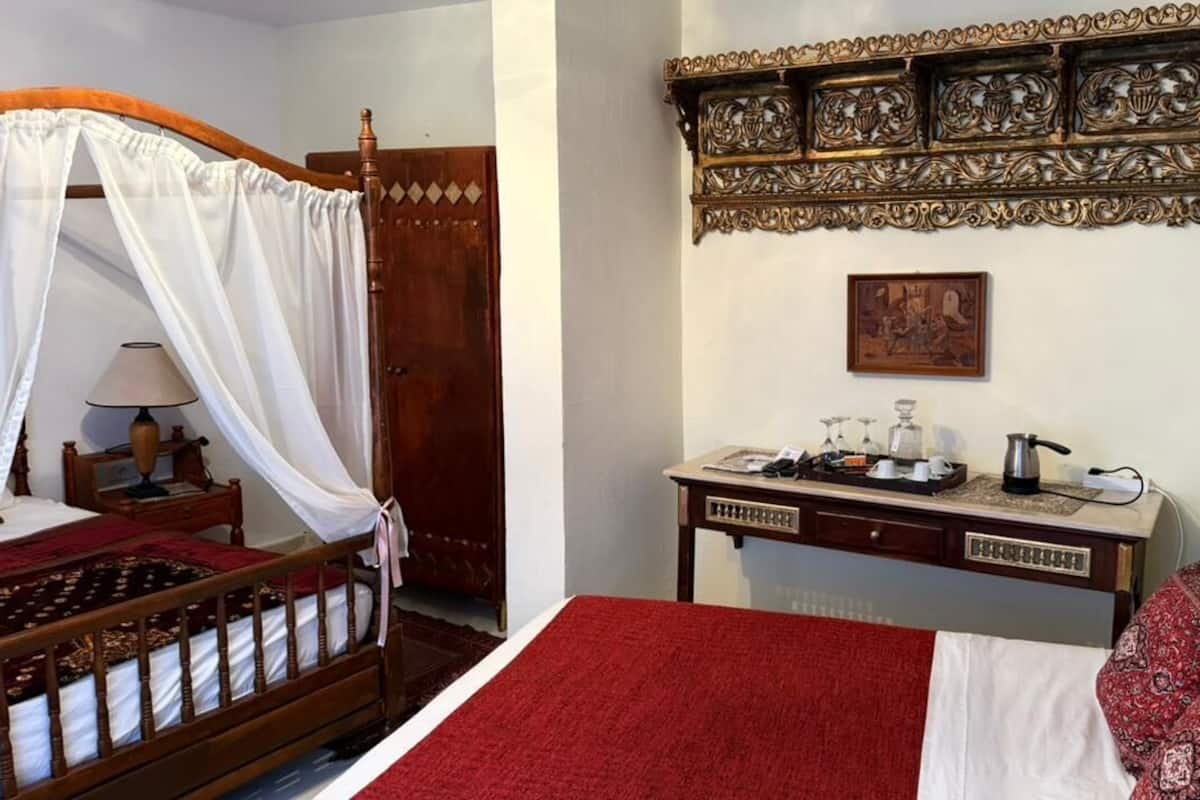 A bedroom with a four-poster bed, a bedside table with a lamp, a red rug, and a decorative wall panel.