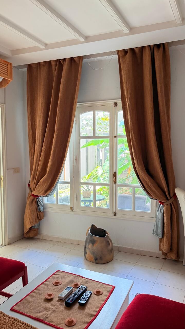 A room with a window, a table with remote controls, a red chair, and a decorative pot.