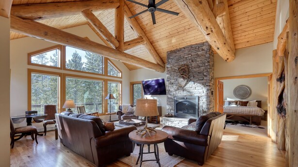 A spacious living room with a stone fireplace, wooden beams, and large windows.
