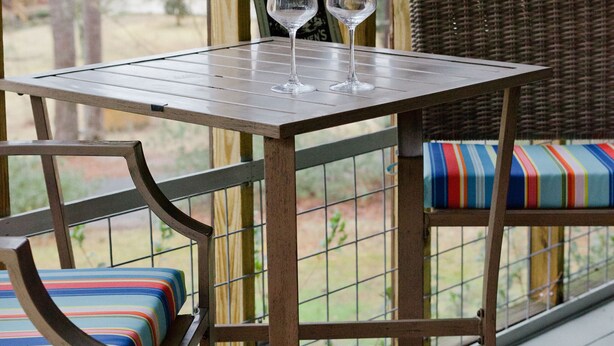 A wooden table with two wine glasses on a deck with wicker furniture and a colorful cushion.