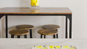 A dining area with a wooden table, two stools, a glass vase with lemons, a clock, and a 'HELLO SUNSHINE' sign.