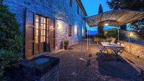 A stone building with wooden doors and windows, a patio with a table and chairs, and a pergola with lights.