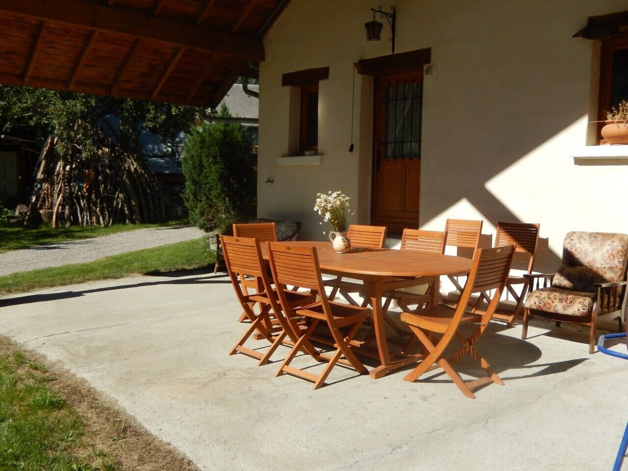 Een terras buiten met een ronde houten tafel en stoelen, een bloempot en een raam met houten kader.