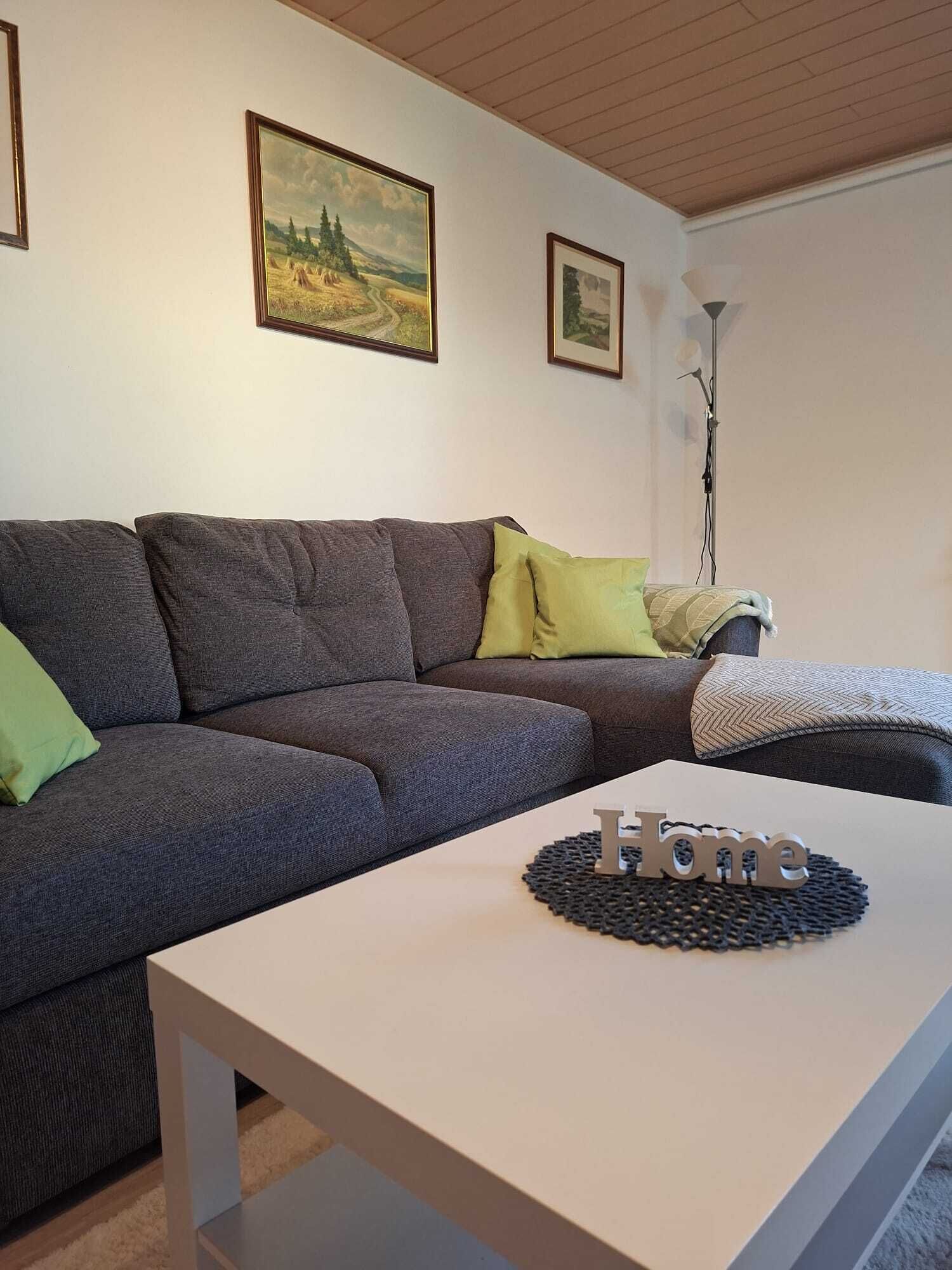 Una sala de estar moderna con un sofá gris, una mesita de centro blanca con un letrero que dice 'Home' y dos cuadros con paisajes enmarcados en la pared.