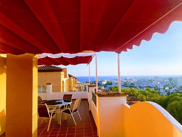 A balcony with a red awning, a table with chairs, and a view of the city and sea.