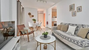 Een moderne woonkamer met een bank, salontafel en stoelen. Links is een televisie te zien en er is een keuken op de achtergrond.