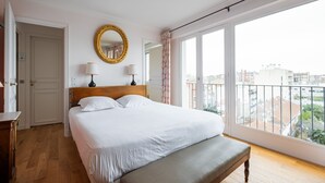 A hotel room with a large bed, a bench, a mirror, two bedside lamps, and a view of the city through large windows.