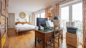 A bedroom with a bed, desk, and computer, large windows with curtains, and a view of the city.
