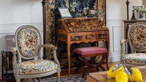 A traditional room with a large tapestry, two upholstered chairs, a wooden side table, and a glass coffee table with yellow decorative items.