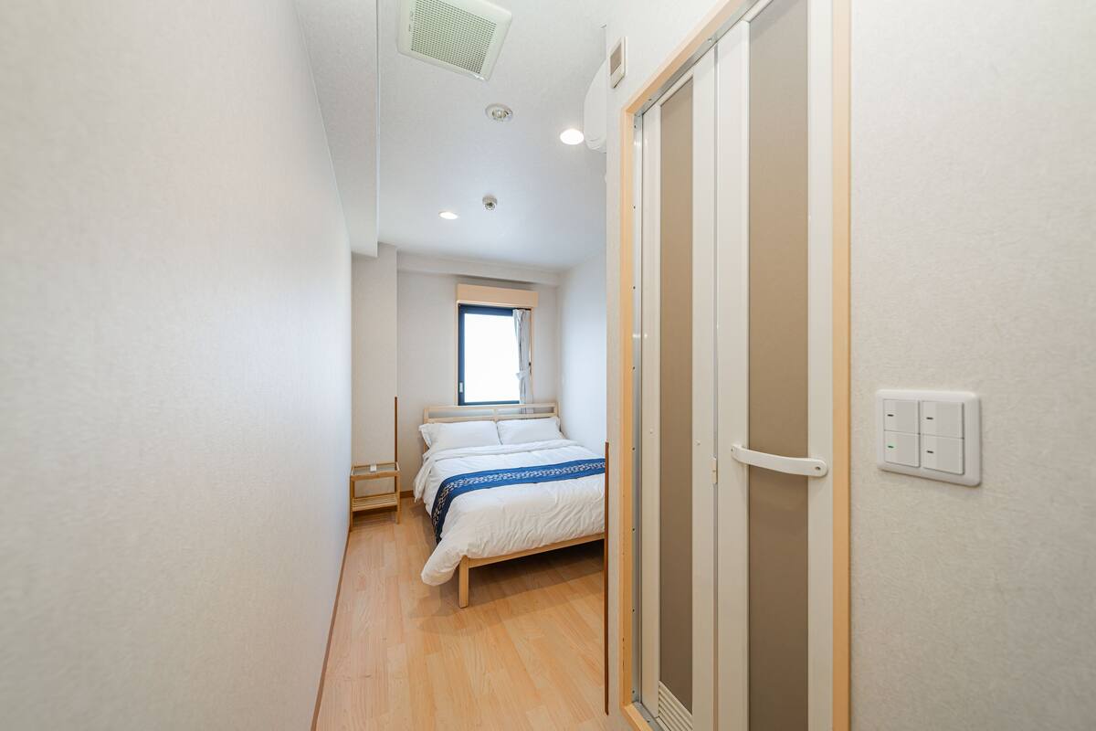 A narrow hallway leading to a bedroom with a bed, a window, and a door.