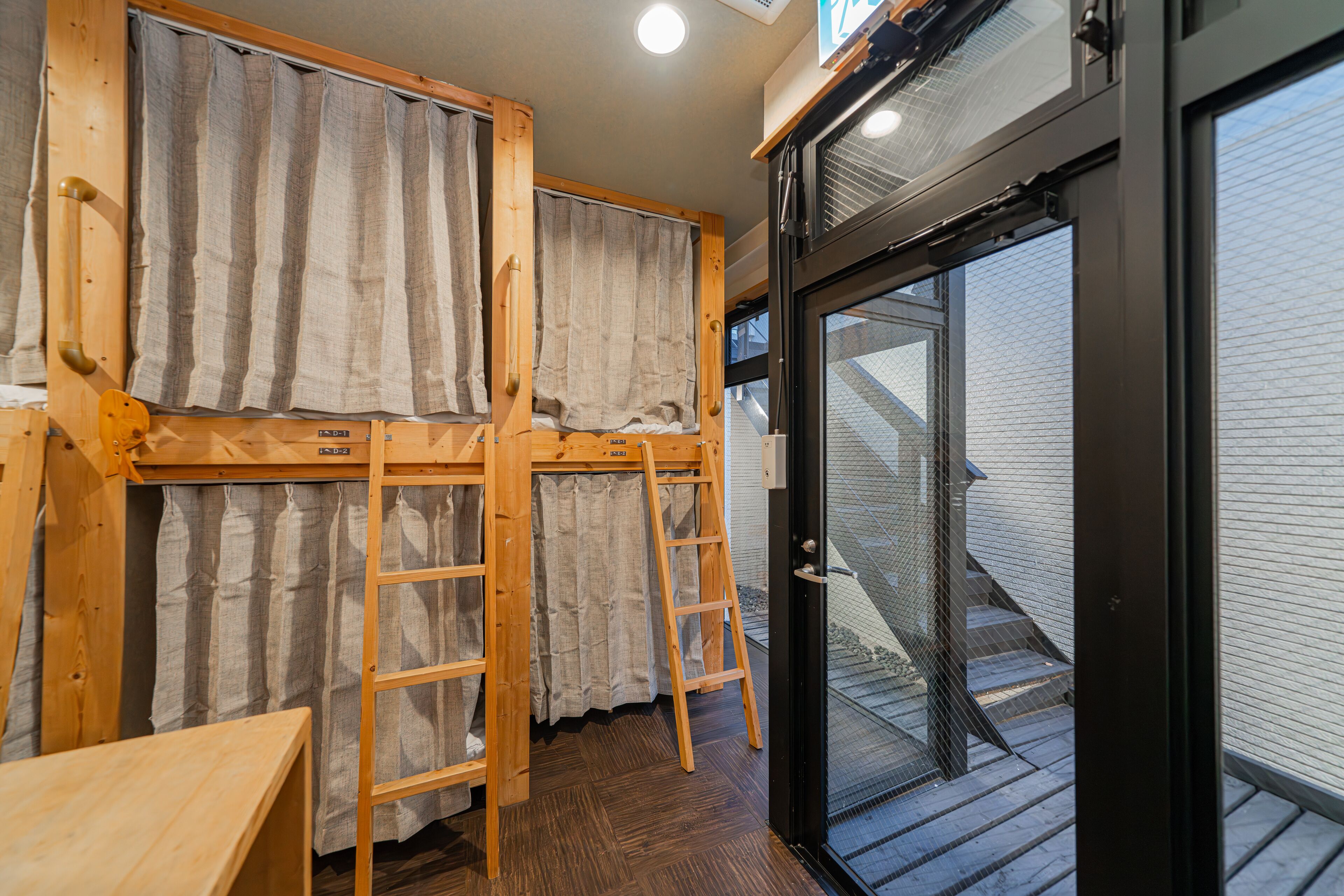 A dormitory room with bunk beds, a wooden ladder, and a sliding glass door leading to a balcony.