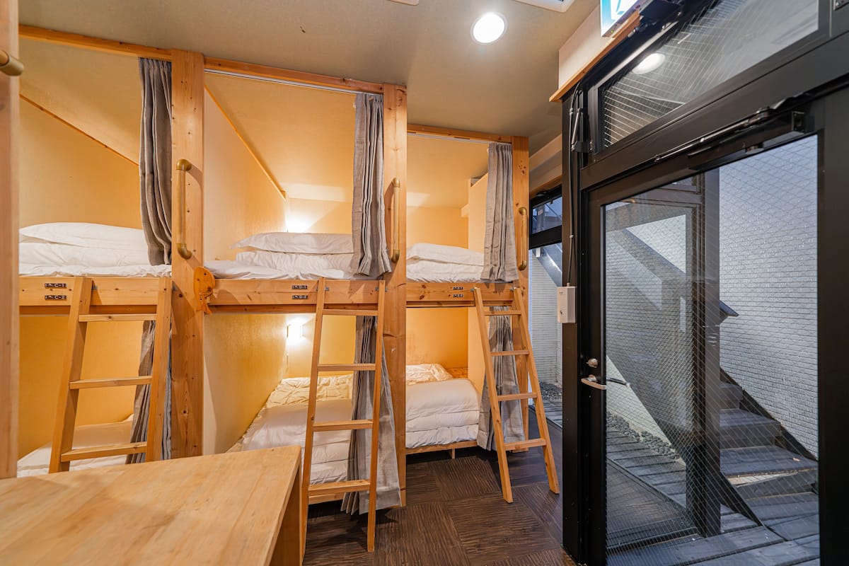 A dormitory room with bunk beds, wooden desks, and a window overlooking a brick building.