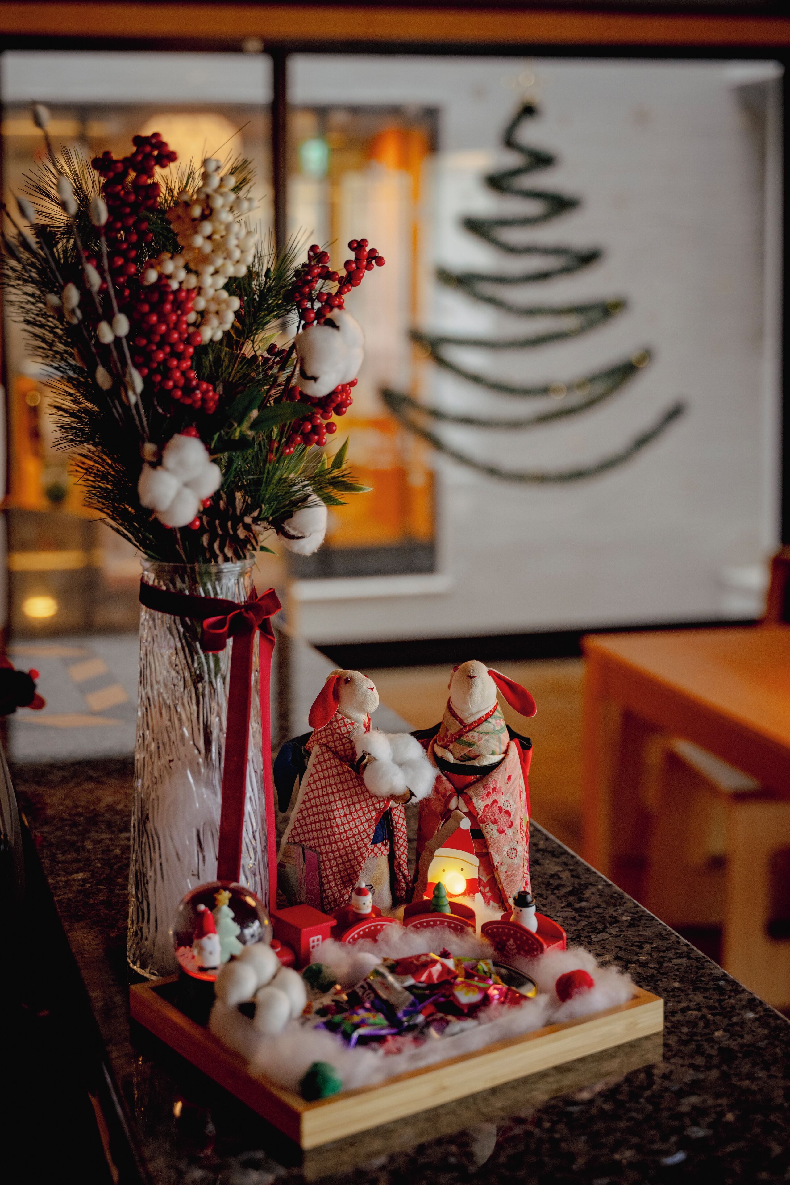 A festive table setting with a vase of greenery and red berries, two small figurines, and a miniature Christmas scene.