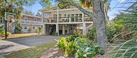 A two-story house with a balcony, surrounded by tropical plants and a basketball hoop.
