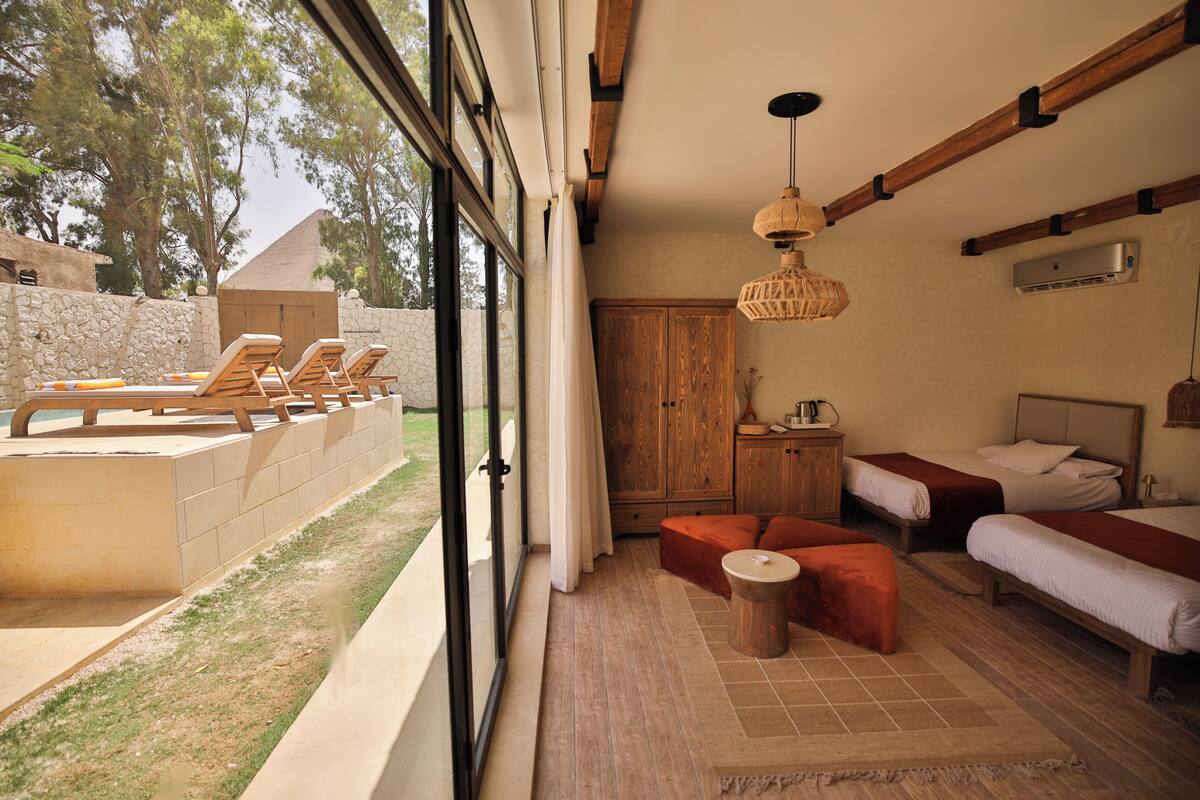 A room with a large window, a wooden ceiling, two beds, a small table, and a stone wall.