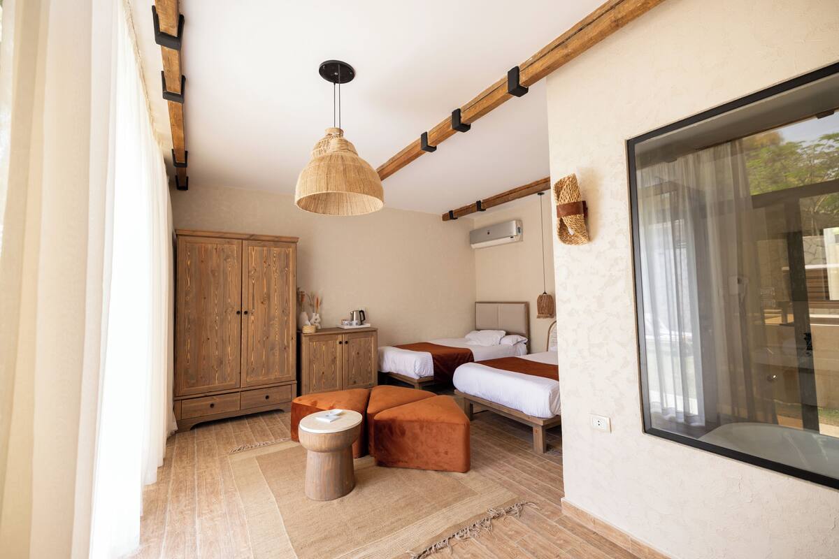 A room with two beds, a wooden wardrobe, a hanging light fixture, and a large window.