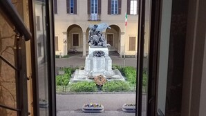 Uitzicht door een deuropening op een binnenplaats met een centraal monument, een gebouw met gebogen ramen en een vlag.