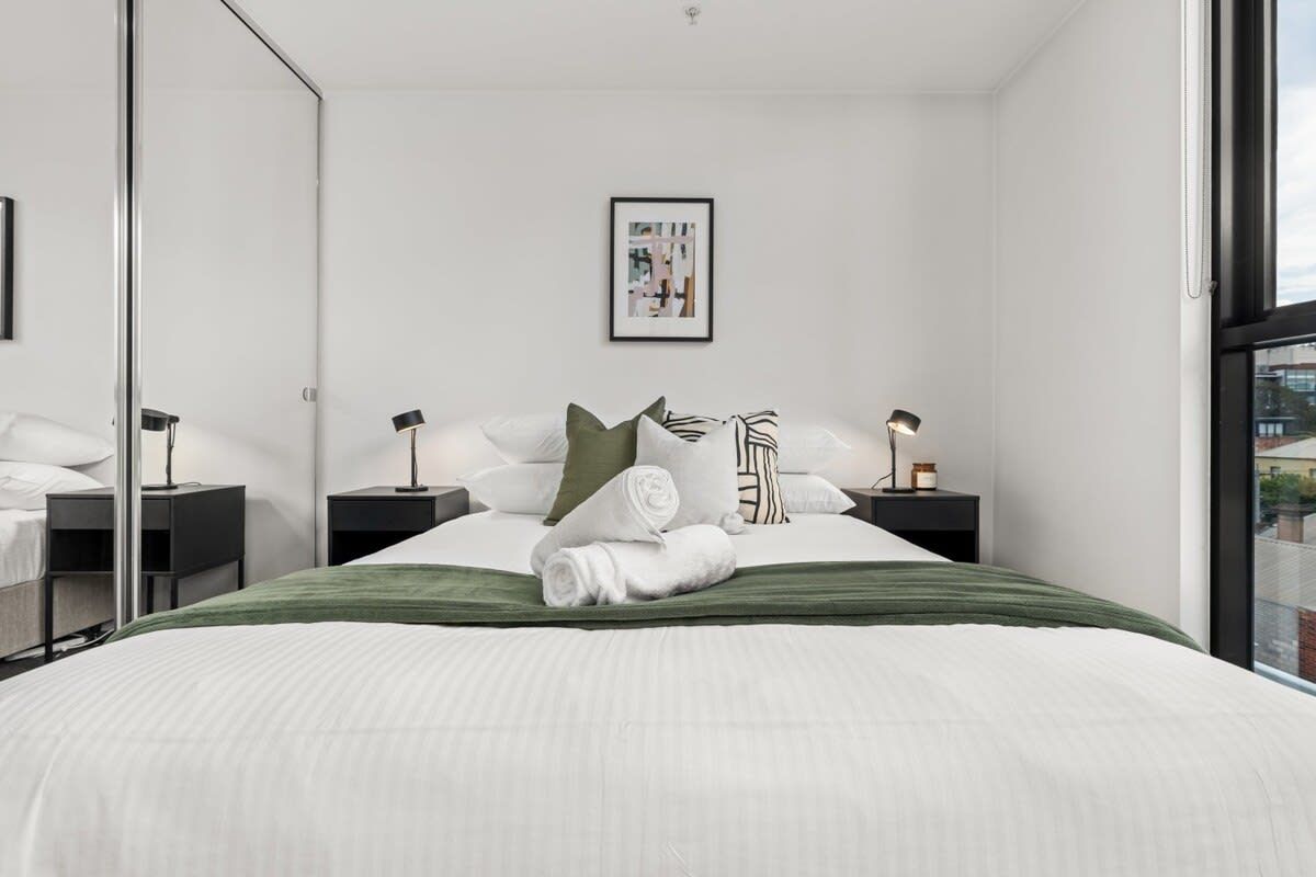 A modern bedroom with a large bed, two bedside tables, a framed picture on the wall, and a view through a window.