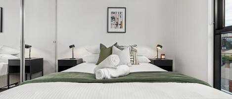 A modern bedroom with a large bed, two bedside tables, a framed picture on the wall, and a view through a window.