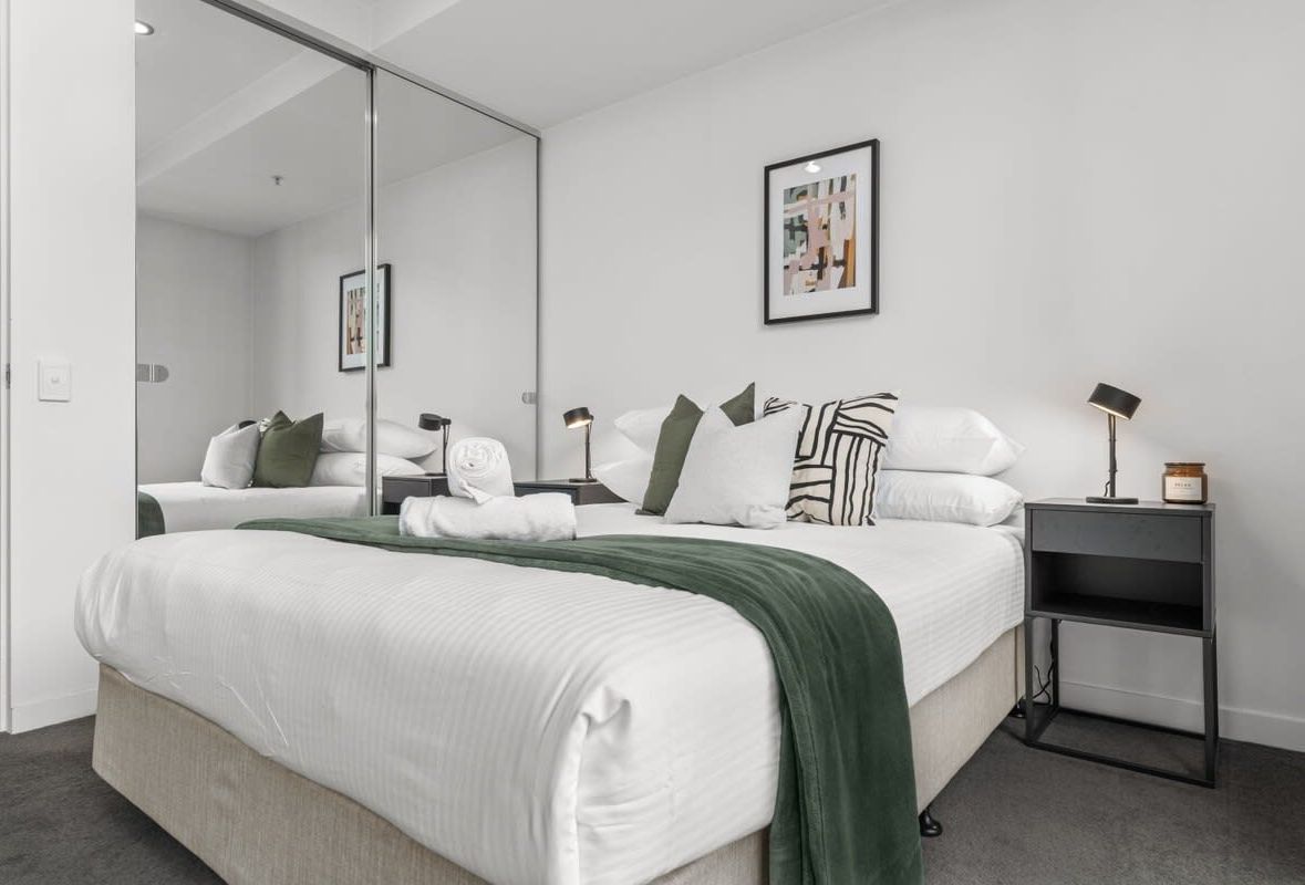 Ein modernes Hotelzimmer mit einem großen Bett, einem Nachttisch mit Lampe, einem Spiegel und einem gerahmten Bild an der Wand.