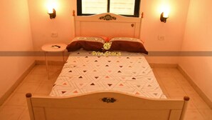 A bedroom with a bed, two wall-mounted lights, a small round table, and a window with blinds. - Privy Stays Reed Field Cottage (Alibag)