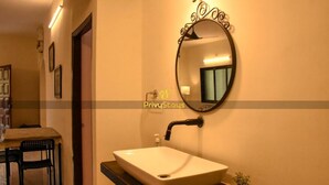 A bathroom with a white sink, a round mirror with a decorative frame, and a small potted plant. - Privy Stays Reed Field Cottage (Alibag)