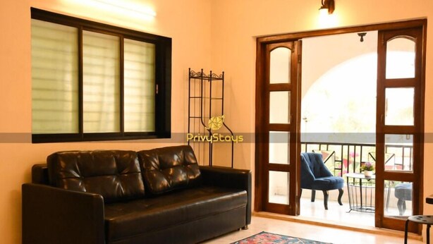 A living room with a black leather sofa, a patterned rug, a wooden door leading to a balcony, and a metal gate. - Privy Stays Reed Field Cottage (Alibag)