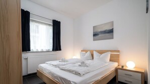 A modern hotel room with a wooden bed, white linens, a bedside table with a lamp, and a view of the city through the window. - Ferienwohnung Uferpromenade mit Terrasse (Röbel/Müritz)