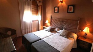Un llit doble amb una capçalera de fusta, dues taules de nit amb làmpades, una cadira i una finestra amb cortines.