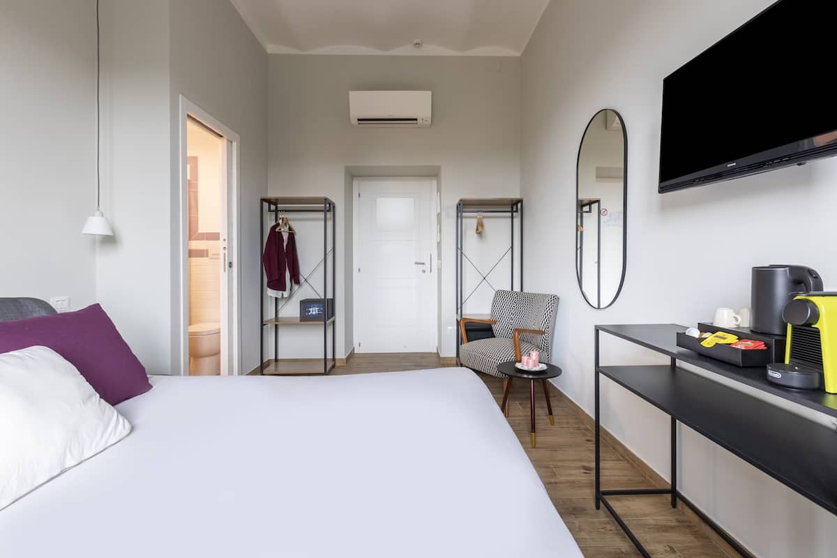 A hotel room with a bed, a desk with a coffee maker, a chair, a wardrobe, and a TV mounted on the wall.