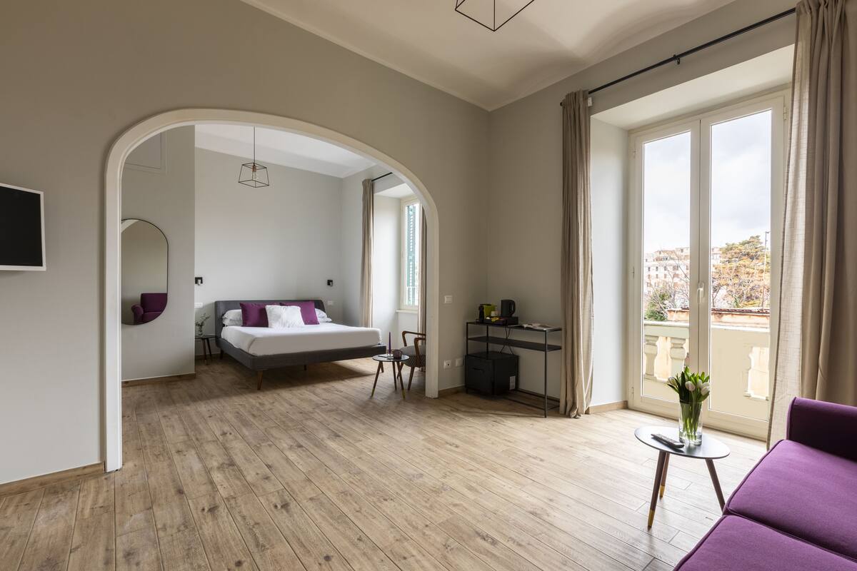 A modern hotel room with a wooden floor, a bed, a desk, and a balcony with a view.