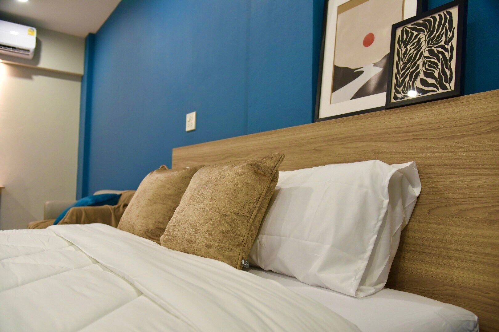 A neatly made bed with white bedding and two beige pillows, a wooden headboard, and a blue wall with two framed artworks.
