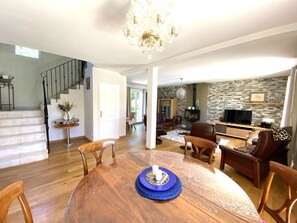 Uma sala de estar ampla com uma mesa de jantar de madeira, poltronas de couro, lareira e televisão.