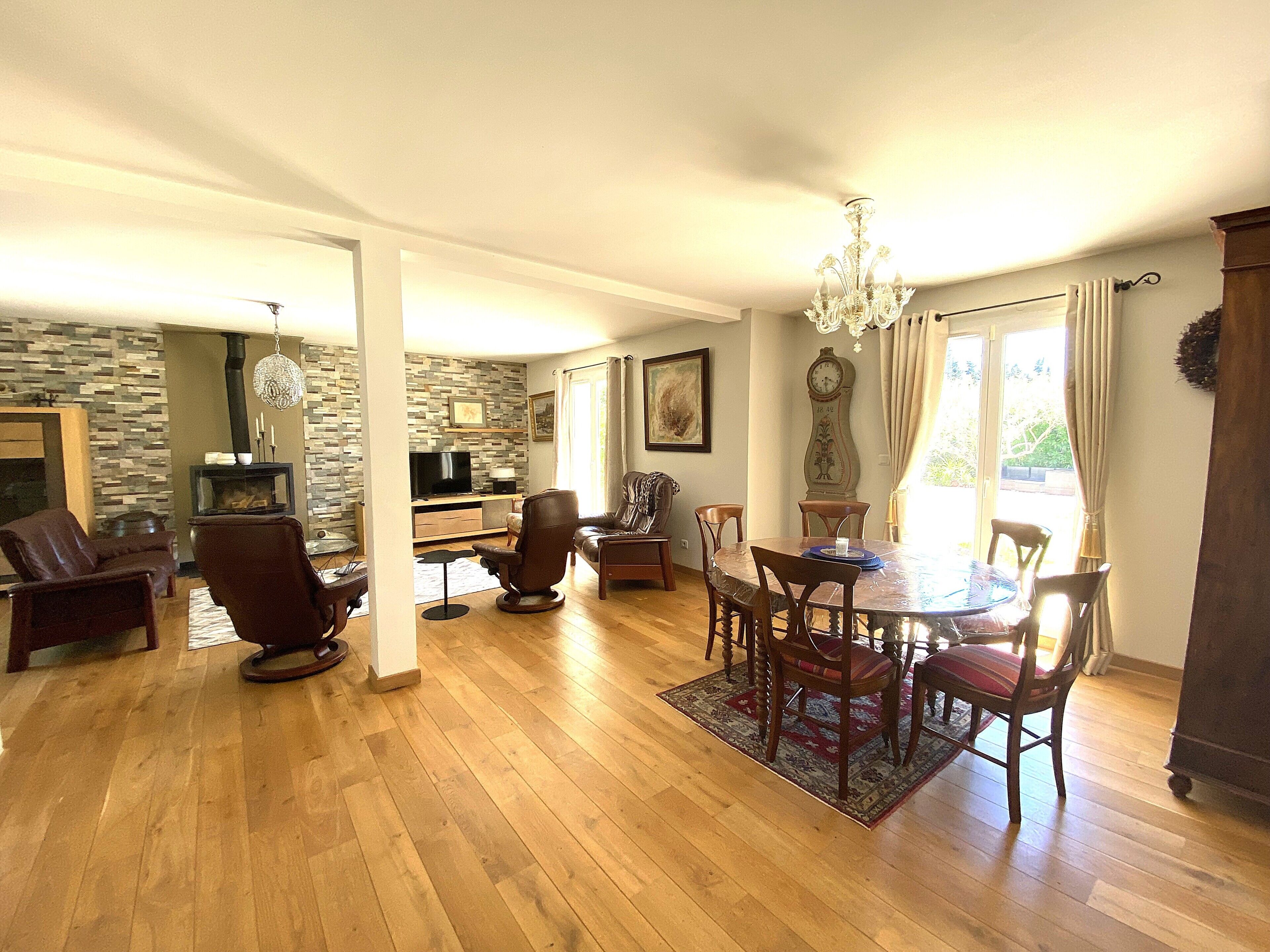 A spacious living area with a fireplace, a dining table with chairs, a chandelier, and a large clock on the wall.