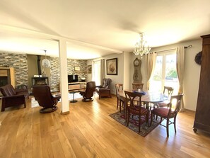 A spacious living area with a fireplace, a dining table with chairs, a chandelier, and a large clock on the wall.