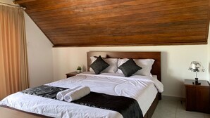 Ein ordentlich bezogenes Bett mit weißen Laken, zwei dunklen Kissen und einer schwarzen Tagesdecke. Dazu ein hölzernes Kopfteil, ein kleiner Nachttisch mit Lampe sowie ein Vorhang links.