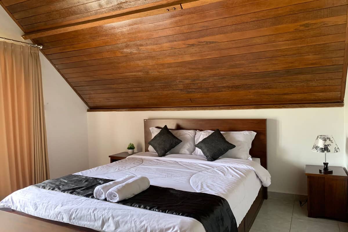 A neatly made bed with white linens, two dark pillows, and a black throw blanket. There is a wooden headboard, a small bedside table with a lamp, and a curtain on the left.