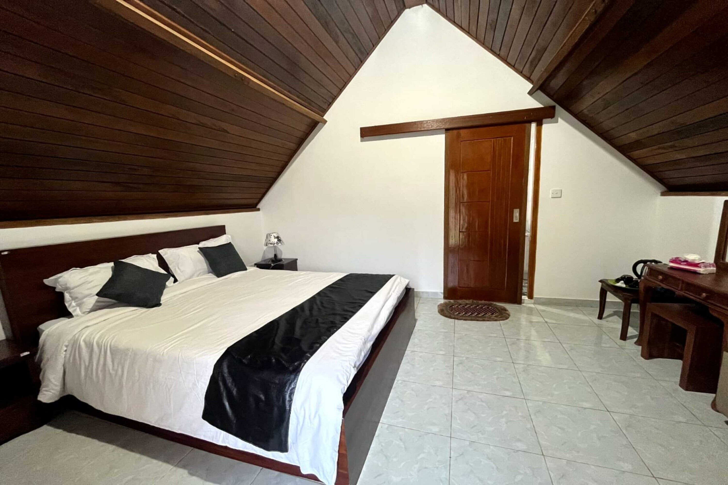 Un dormitorio con techo de madera, una cama con ropa de cama blanca y negra, un escritorio de madera y una puerta.