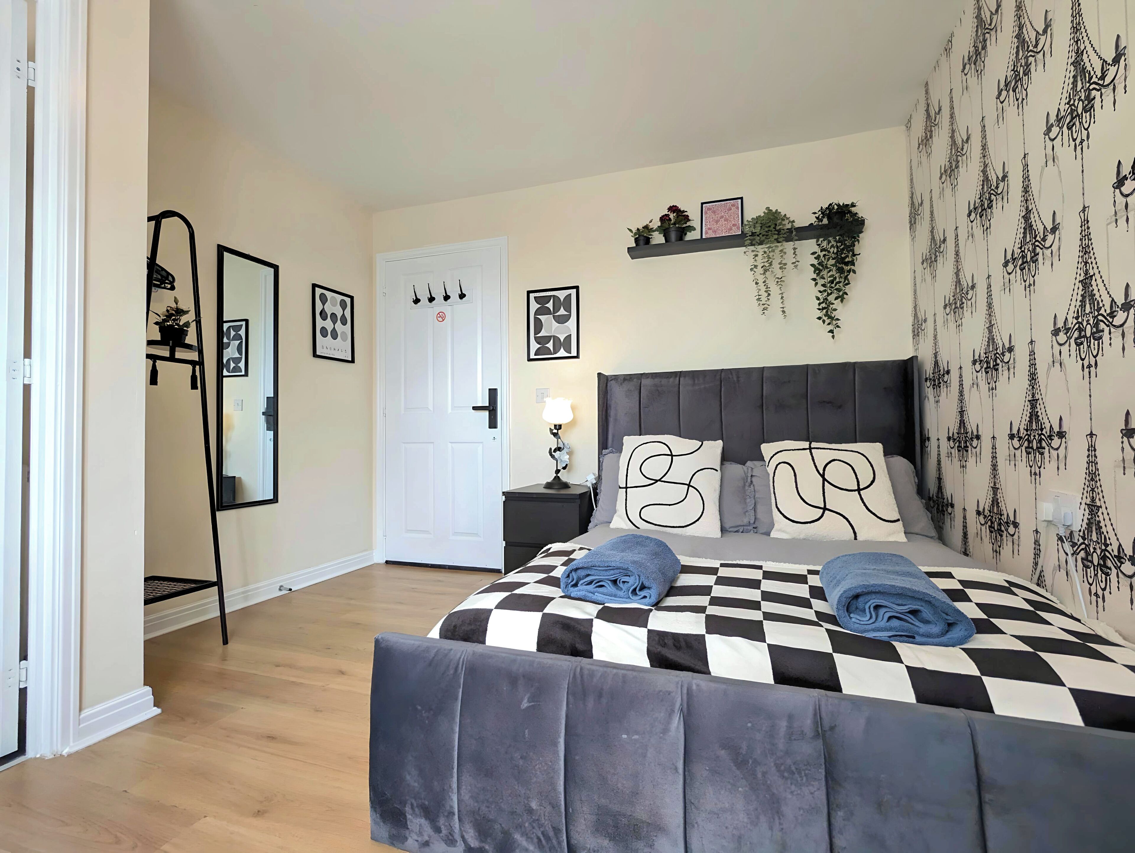Ein Schlafzimmer mit kariertem Bettbezug, einem grau gepolsterten Kopfteil und einem an der Wand montierten Regal mit Pflanzen und Bildern.