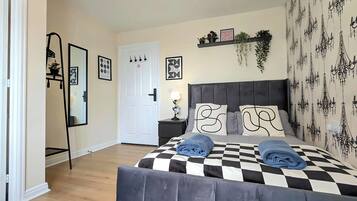 Ein Schlafzimmer mit kariertem Bettbezug, einem grau gepolsterten Kopfteil und einem an der Wand montierten Regal mit Pflanzen und Bildern.